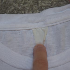 Op de afbeelding: Close-up van een witte t-shirtkraag met een label. De kraag is geribbeld en het label heeft een lichtcr&egrave;mekleur. Het shirt lijkt gemaakt van een zacht, lichtgewicht materiaal.