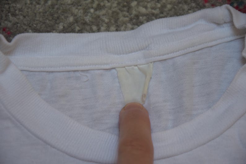 Op de afbeelding: Close-up van een witte t-shirtkraag met een label. De kraag is geribbeld en het label heeft een lichtcr&egrave;mekleur. Het shirt lijkt gemaakt van een zacht, lichtgewicht materiaal.
