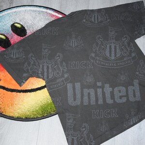 K&ouml;nnte beinhalten: Ein schwarzes T-Shirt mit einem sich wiederholenden Muster des Newcastle United-Wappens und dem Wort "Kick" in grauem Druck.