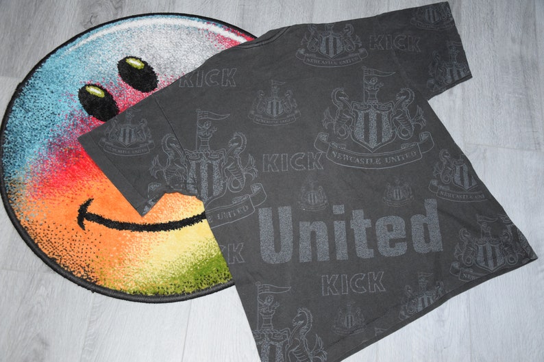 K&ouml;nnte beinhalten: Ein schwarzes T-Shirt mit einem sich wiederholenden Muster des Newcastle United-Wappens und dem Wort "Kick" in grauem Druck.