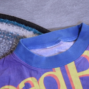 Puede incluir: Una camiseta morada con un estampado gr&aacute;fico amarillo y rojo que dice "Good Vibes". La camiseta tiene un cuello de canal&eacute; azul.