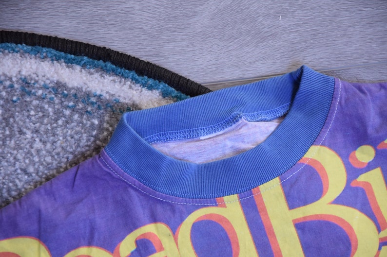 Puede incluir: Una camiseta morada con un estampado gr&aacute;fico amarillo y rojo que dice "Good Vibes". La camiseta tiene un cuello de canal&eacute; azul.
