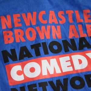 K&ouml;nnte beinhalten: Blaues T-Shirt mit dem Aufdruck "NEWCASTLE BROWN ALE NATIONAL COMEDY NETWORK" in Rot, Schwarz und Wei&szlig;. Der Text ist in einer gestapelten Anordnung angeordnet, wobei jede Textzeile &uuml;bereinander liegt.