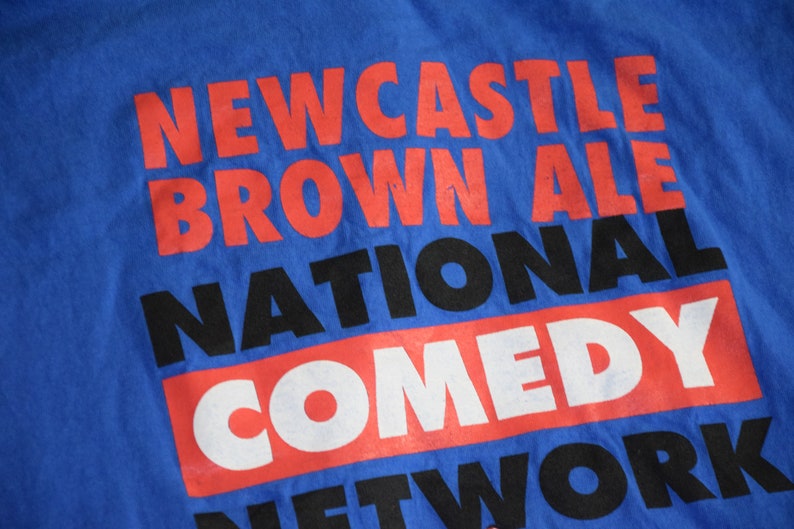 K&ouml;nnte beinhalten: Blaues T-Shirt mit dem Aufdruck "NEWCASTLE BROWN ALE NATIONAL COMEDY NETWORK" in Rot, Schwarz und Wei&szlig;. Der Text ist in einer gestapelten Anordnung angeordnet, wobei jede Textzeile &uuml;bereinander liegt.