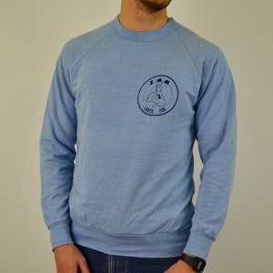 Puede incluir: Una sudadera azul claro con un logotipo circular azul marino que presenta el texto "E.M.M. Lakes 1985" y un gráfico de un lago.