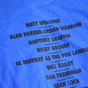 K&ouml;nnte beinhalten: Nahaufnahme eines blauen T-Shirts mit schwarzem Text. Der Text enth&auml;lt Namen wie "MATT WELCOME", "ALAN PARKER-URBAN WARRIOR" und "AL MURRAY AS THE PUB LANDLORD". Das Shirt besteht aus weichem Stoff.