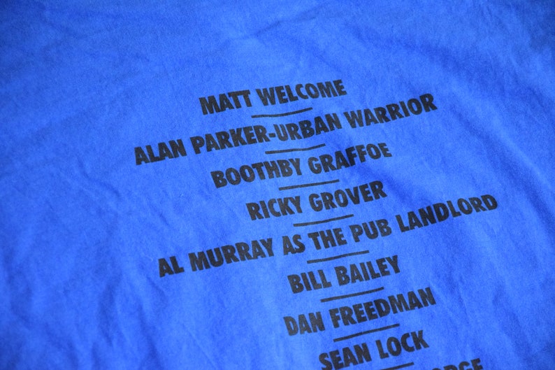 K&ouml;nnte beinhalten: Nahaufnahme eines blauen T-Shirts mit schwarzem Text. Der Text enth&auml;lt Namen wie "MATT WELCOME", "ALAN PARKER-URBAN WARRIOR" und "AL MURRAY AS THE PUB LANDLORD". Das Shirt besteht aus weichem Stoff.