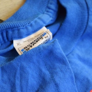 K&ouml;nnte beinhalten: Nahaufnahme eines blauen T-Shirts mit einem Etikett von Screen Stars by Fruit of the Loom. Das Etikett weist die Gr&ouml;&szlig;e XL aus. Das T-Shirt besteht aus 100 % Baumwolle.