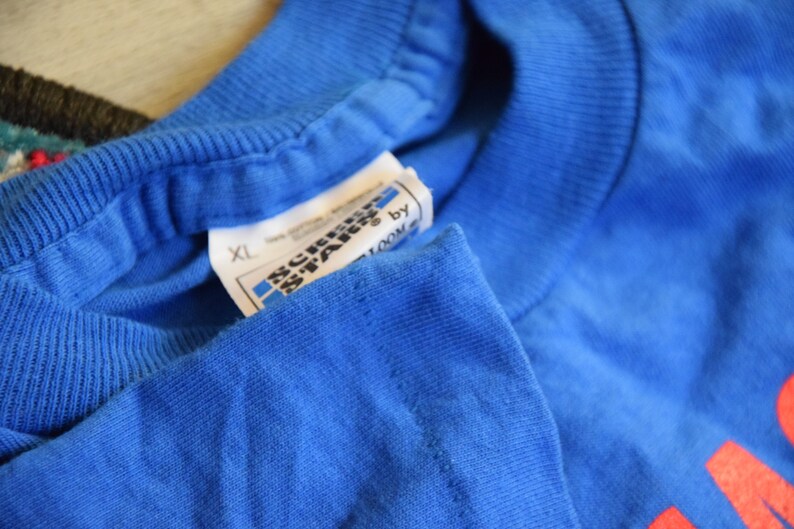 K&ouml;nnte beinhalten: Nahaufnahme eines blauen T-Shirts mit einem Etikett von Screen Stars by Fruit of the Loom. Das Etikett weist die Gr&ouml;&szlig;e XL aus. Das T-Shirt besteht aus 100 % Baumwolle.