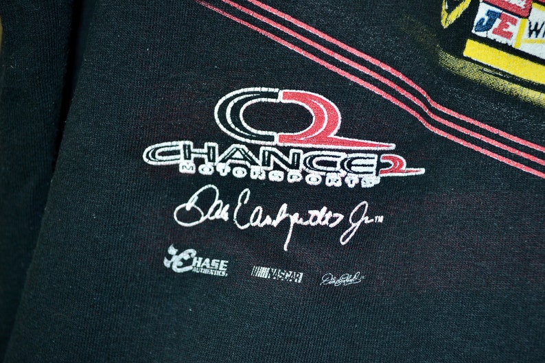 Op de afbeelding: Zwart t-shirt met het Chance 2 Motorsports logo in wit en rood, en de handtekening van Dale Earnhardt Jr. Het shirt toont ook de logo's van Chase Authentics en NASCAR.