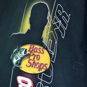 Op de afbeelding: Zwart t-shirt met het Bass Pro Shops-logo, een visafbeelding op een gele ovaal en witte tekst. Een silhouetontwerp en het nummer 8 zijn ook zichtbaar. Het t-shirt is gemaakt van zacht materiaal.
