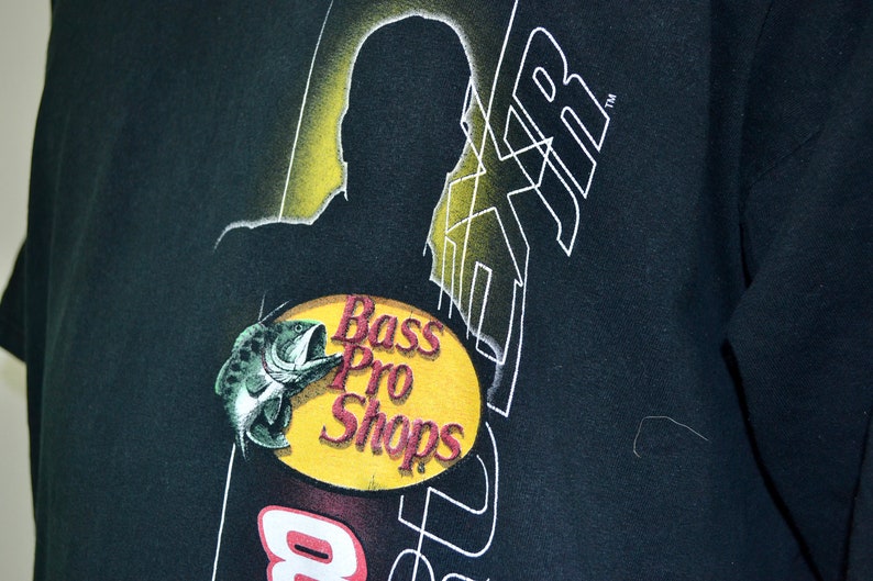 Op de afbeelding: Zwart t-shirt met het Bass Pro Shops-logo, een visafbeelding op een gele ovaal en witte tekst. Een silhouetontwerp en het nummer 8 zijn ook zichtbaar. Het t-shirt is gemaakt van zacht materiaal.
