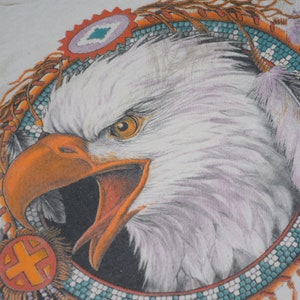 May include: A white bald eagle with a bright orange beak and yellow eyes is depicted in a Native American headdress. The headdress is made of feathers, beads, and a beaded band with turquoise and orange accents.