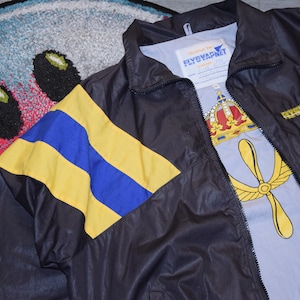 May include: A black nylon jacket with yellow and blue stripes on the left sleeve. The jacket has a zipper closure and a label that reads "Flygvapnet" on the inside of the collar.