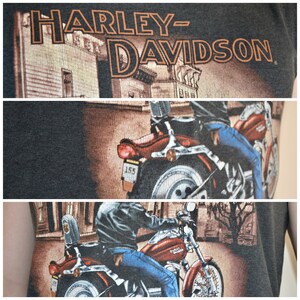May include: Dark gray Harley-Davidson t-shirt featuring a motorcycle graphic. The graphic shows a person riding a red motorcycle with the Harley-Davidson logo. The shirt has the Harley-Davidson logo printed across the top.