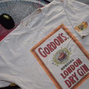 May include: Cream-colored t-shirt featuring a large rectangular graphic with the text "Gordon's Special London Dry Gin." The graphic includes a fox illustration and a red, orange, and white border. A small logo is on the sleeve.