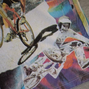 Puede incluir: Una camiseta colorida con un estampado gr&aacute;fico de dos personas montando en bicicleta. La impresi&oacute;n presenta un fondo de arco&iacute;ris con salpicaduras de color.
