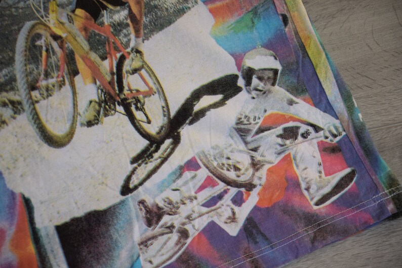 Puede incluir: Una camiseta colorida con un estampado gr&aacute;fico de dos personas montando en bicicleta. La impresi&oacute;n presenta un fondo de arco&iacute;ris con salpicaduras de color.