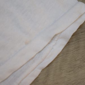 Op de afbeelding: Close-up van gevouwen witte stof, mogelijk een t-shirt of ander kledingstuk. De stof is licht gekreukt en heeft een zachte textuur. De rand is omgeslagen en toont de naad en de dikte van het materiaal.