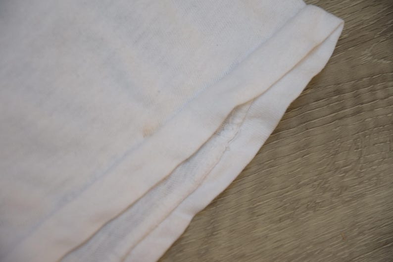 Op de afbeelding: Close-up van gevouwen witte stof, mogelijk een t-shirt of ander kledingstuk. De stof is licht gekreukt en heeft een zachte textuur. De rand is omgeslagen en toont de naad en de dikte van het materiaal.