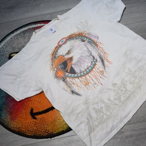 May include: White t-shirt with a graphic print of a bald eagle in a Native American dreamcatcher design. The eagle is facing forward with its beak open and has a white head and brown body. The dreamcatcher is made of feathers and beads.