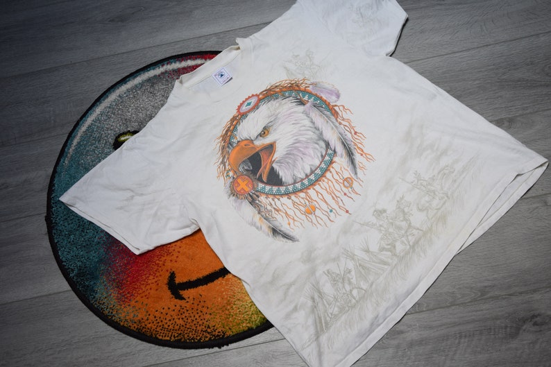 May include: White t-shirt with a graphic print of a bald eagle in a Native American dreamcatcher design. The eagle is facing forward with its beak open and has a white head and brown body. The dreamcatcher is made of feathers and beads.