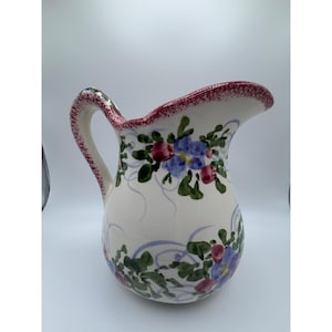 May include: A ceramic pitcher with a cream-colored base, featuring hand-painted blue flowers, green leaves, and small red berries. The rim and the handle are decorated with a speckled, dark red trim, and light purple swirls accent the floral design.