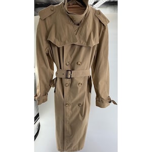 May include: A tan double-breasted trench coat featuring a wide collar, shoulder epaulets, and a matching waist belt with a rectangular buckle. The coat includes long sleeves with adjustable cuff straps, a storm flap, and two rows of buttons.