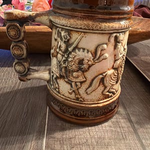 May include: A vintage ceramic beer stein with a brown glaze and detailed relief carvings. The handle features decorative circular accents. The stein depicts figures on horseback and other medieval scenes, showcasing a rustic, antique aesthetic.