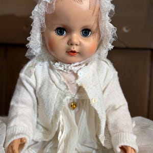 May include: A vintage doll with blue eyes, rosy cheeks, and red lips, wearing a white lace bonnet, a cream-coloured knit cardigan with buttons, and a sheer white dress. A small gold-toned necklace with a yellow heart-shaped pendant is worn.