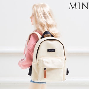 May include: A cream-coloured backpack with a front pocket and a small rectangular label that reads "MINISH". The backpack has black straps and a brown leather zip pull. The word "MINISH" is also printed in the upper right corner.