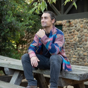 Male model sitting on bench wearing a cardigan with blue, red, and turquoise accents.
