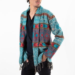 Man wearing stylish long-sleeved cardigan with a t-shirt underneath. Cardigan has geometric patterns with fringe hanging on the ends. Cardigan is turquoise, red, and white.