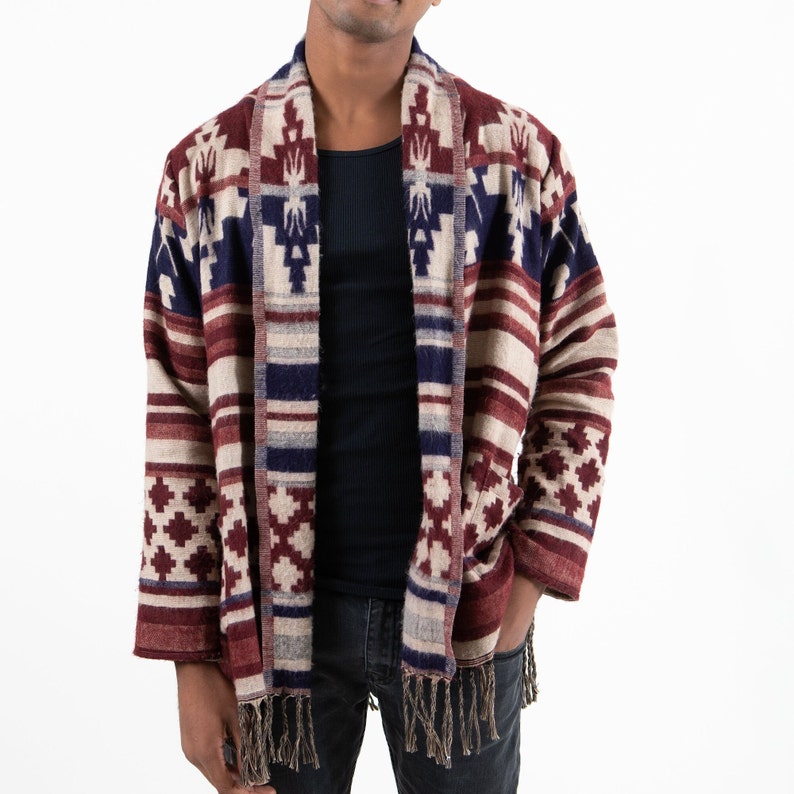 Man wearing a long-sleeved cardigan. Cardigan has geometric patterns with fringe hanging on the ends. Cardigan is red, white, and blue.