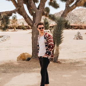 Male modeling in desert in a cardigan with fringes on the ends. Cardigan is burgundy with cream and navy blue accents.