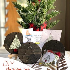 May include: A DIY Christmas tree string art kit featuring three circular dark wood plaques. One plaque shows a completed tree with green string, one has silver nails arranged in a tree shape, and one includes a plastic bag with string and a pattern.