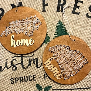 May include: Two circular wooden ornaments featuring string art maps of North Carolina and South Carolina. Each ornament has a white string outline of the state, silver nail accents, and the word home in a light-colored script font with a white hanging cord.