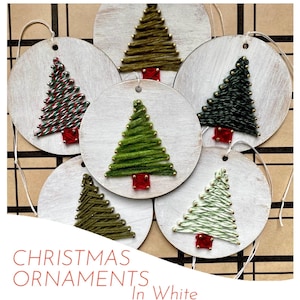 May include: A collection of six white wooden Christmas ornaments. Each ornament features a stitched Christmas tree design in various shades of green, red, and white. The text "CHRISTMAS ORNAMENTS In White" is at the bottom.