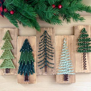 May include: Five rectangular wooden plaques featuring string art designs of Christmas trees. Each tree is created with colored thread wrapped around small nails on a stained wood board, showcasing various shades of green and white thread.