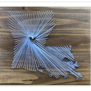 May include: A white string art piece in the shape of the state of Louisiana with a heart in the center. The art is on a wooden background.