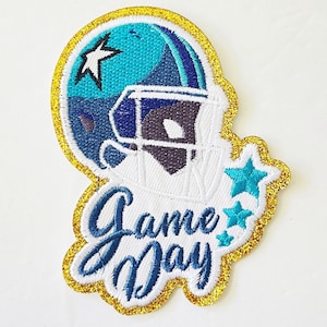 May include: Embroidered patch featuring a football helmet in shades of blue and white, with a gold glitter border. The text "Game Day" is in a script font. Includes star accents.