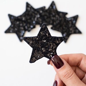 May include: A collection of five-pointed star-shaped patches covered in small, reflective black sequins. Each star features a dark fabric border, and the sequins create a textured, shimmering surface that catches the light across the entire star shape.