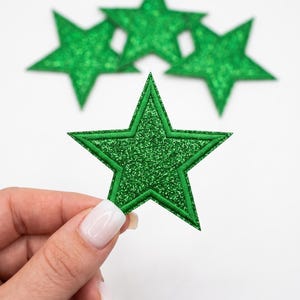 May include: Four five-pointed star-shaped patches featuring a vibrant green glitter finish. Each star has a stitched border detail around the edges. The patches are arranged with one in the foreground and three slightly out of focus behind it.