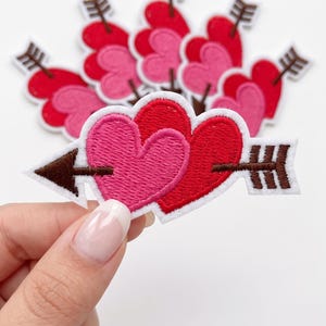 May include: An embroidered patch featuring two overlapping hearts, one pink and one red, pierced by a brown arrow with a white border. The design includes a pointed arrowhead on the left and fletching on the right, with multiple identical patches.