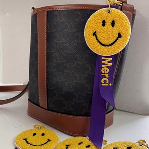 May include: Four yellow smiley face keychains with purple ribbon straps that say "Merci" in gold lettering. The keychains are made of fuzzy fabric and have black embroidered eyes and mouths.