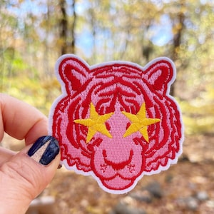 May include: Pink embroidered iron-on patch of a tiger with yellow star-shaped eyes.