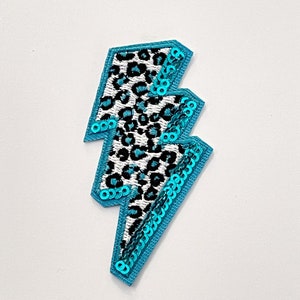 May include: A turquoise lightning bolt shaped patch with a black and white leopard print design. The patch is covered in sequins.
