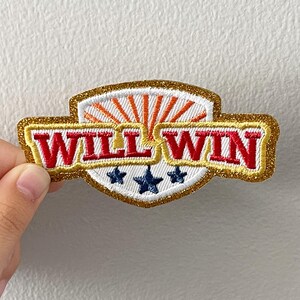 May include: Embroidered patch with the words "WILL WIN" in red, outlined in gold glitter. The patch features a white shield design with orange sun rays and three blue stars. The patch is held by a hand.
