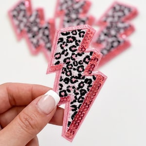 May include: A lightning bolt shaped patch featuring a black and white leopard print pattern on the main body, bordered by a pink stitched edge and accented with a vertical row of shiny pink sequins along the right side of the bolt shape.