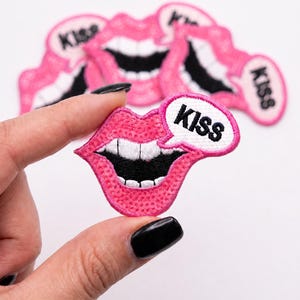 May include: An embroidered patch featuring a pair of bright pink lips with white teeth and a black mouth interior. A white speech bubble attached to the side contains the word KISS in bold black capital letters. The patch has a pink border.
